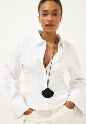 Mujer con una camisa blanca texturizada abotonada y pantalones crema, con las manos en los bolsillos y un collar largo negro con un colgante de concha.