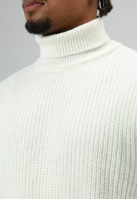 Close-up van een persoon die een wit gebreide coltrui draagt, met getextureerde stof en een deel van het onderste gezicht en een baard zichtbaar.