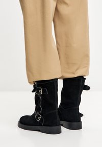 Crne čizme od brušene kože do pola lista imaju bočni patentni zatvarač, dvije kopče i ravni potplat. Čizme imaju teksturiranu završnu obradu i jednostavan dizajn.