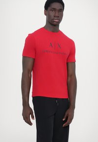 Raudona medvilninė t-shirt su trumpomis rankovėmis, apvaliu kaklu ir ekranu atspausdintu juodu logotipu "A|X ARMANI EXCHANGE" per krūtinę. Juodos kelnės.