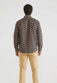 Chemise à manches longues marron foncé avec un motif géométrique crème et rouge. Dotée d'un col ouvert et de poignets à boutons. Portée avec un pantalon beige clair.