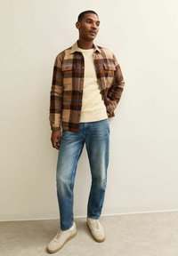 Geruite bruine en blauwe flanellen shirt over een crèmekleurige gebreide trui, gecombineerd met lichtblauwe jeans en beige sneakers, staand tegen een neutrale muur.