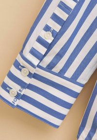 Blue and white striped shirt cuff with two buttons, featuring contrasting stitching and embroidered brand name. Smooth cotton texture.