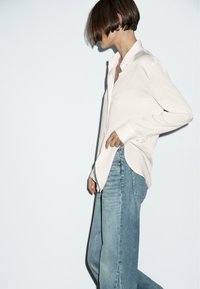 White long-sleeve shirt with a buttoned placket and curved hem, paired with light blue high-waisted jeans in a relaxed fit.