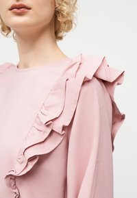 Γυναίκα με σγουρά ξανθά μαλλιά που φορεί ένα ανοιχτό ροζ μπλουζάκι με έντονη λεπτομέρεια βολάν στους ώμους και το στήθος