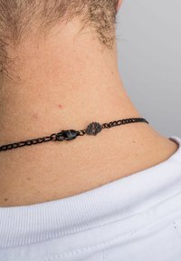 Melnā ķēdes kaklarota ar sprādzi, kas rotāta ar dekoratīvu emblemu. Ķēde ir eleganta ar nelielu spīdumu uz gaišas ādas fona.