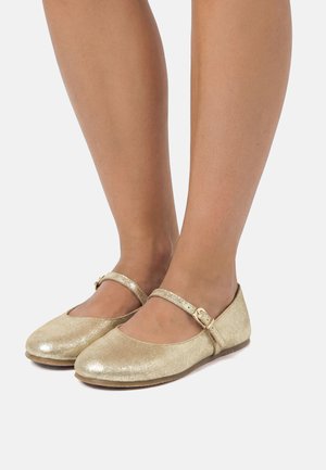 Füße, die metallisch goldene Mary Jane Ballerinas mit Schnallenriemen tragen, vor einem einfarbigen weißen Hintergrund gezeigt.