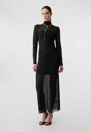 Femme portant une robe longue noire en voile à manches longues avec de petits points blancs, un collier doré à plusieurs rangs, et des escarpins noirs ouverts à l'avant.