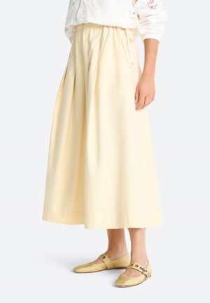 Frau trägt eine hoch taillierte, weite kremfarbene Hose mit Falten, kombiniert mit blassgelben flachen Schuhen mit Riemen und Metallösen.