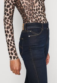 Tunna mörkblå denimjeans med kontrasterande gula sömmar, försedda med framfickor och en etikett på baksidan av midjebandet.