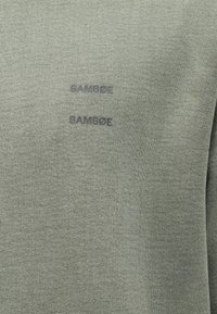 Pogled od blizu na zeleni pleteni material z dvema rahlo dvignjenima logoma "SAMSOE" v temno sivi barvi.