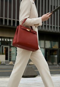 Osoba u krem odijelu hoda na otvorenom, držeći tablet s velikom crvenom kožnom torbom na ruci.