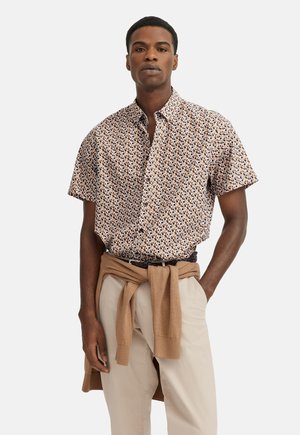 Kurzarm-Hemd mit Knöpfen in einem blassbraunen, blauen und dunkelbraunen Blumenmuster; getragen mit beigen Hosen und einem braunen Pullover, der um die Taille gebunden ist.