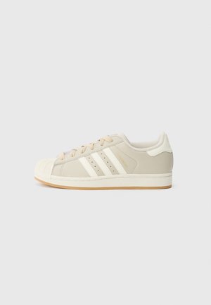 Beige lavt snøret sneaker med hvid gummiskal på tåen, tre hvide sidelister, beige snørebånd og en gummisål på en lys grå baggrund.