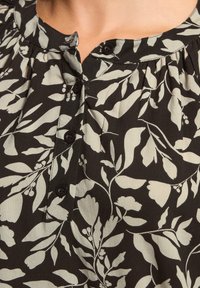Schwarze Bluse mit beigefarbenem Blumenmuster, mit rundem Halsausschnitt und vier schwarzen Knöpfen auf der Vorderseite.