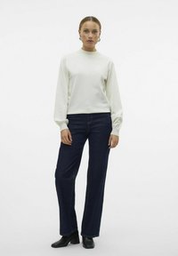 Pull crème à col haut et manches bouffantes, associé à un jean droit bleu marine et des bottines noires.