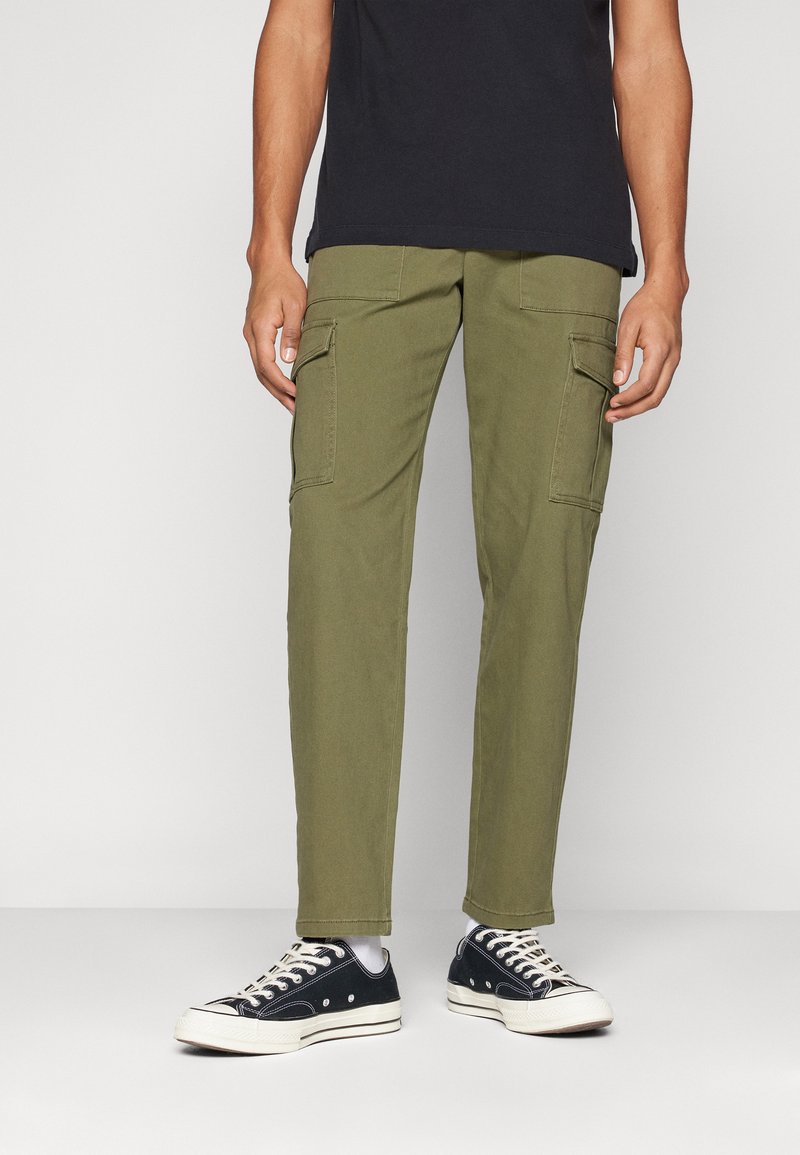 Los pantalones cargo verde olivo cuentan con dos bolsillos laterales, un corte recto y una tela ligera, combinados con zapatillas de lona negras.