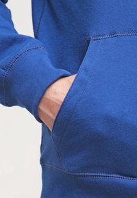 Mano parzialmente dentro della tasca anteriore di una felpa blu a maniche lunghe con dettagli di cucitura visibili.