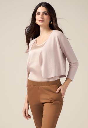 Cardigan court rose clair sur un haut beige, associé à un pantalon beige ajusté ; texture douce, design simple, manches trois-quarts.