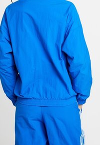 Blå lättviktsjacka med elastiska ärmslut och midjeband, med en slät yta och en avslappnad passform. Inkluderar matchande blå byxor.