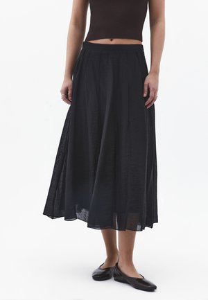 Frau trägt ein schwarzes ärmelloses bauchfreies Top, einen schwarzen langen plissierten Rock und schwarze Ballerinas und steht vor einem weißen Hintergrund.