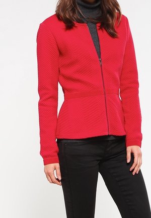 Kobieta w dopasowanej czerwonej kurtce zapinanej na zamek z teksturowanym wzorem w jodełkę, nosząca pod spodem ciemny golf, w połączeniu z czarnymi spodniami.