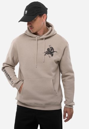Homme portant un sweat à capuche beige avec un motif de bélier sur la poitrine et un texte sur la manche, une casquette noire avec un logo, et un pantalon noir, debout avec la main dans la poche.