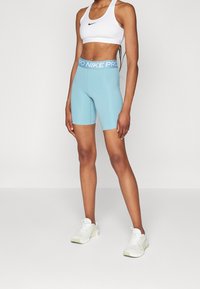 Jasnoniebieskie szorty kompresyjne z szerokim, niebieskim paskiem oznaczonym "PRO NIKE". Noszone z białym stanikiem sportowym i białymi butami sportowymi.