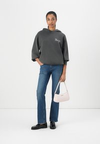 Haut de sweat gris surdimensionné avec logo brodé, jean droit bleu, et sac à main rose clair. Le modèle se tient sur un fond blanc.
