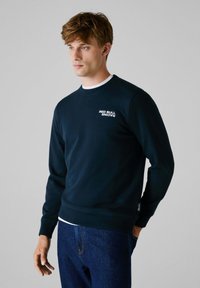 Marineblaues Sweatshirt mit Rundhalsausschnitt, weißem "RED BULL RACING"-Schriftzug auf der Brust und gerippten Bündchen. Kombiniert mit dunklen Jeans.