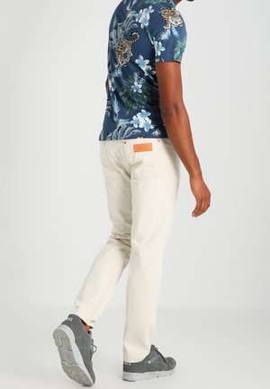 Mies kävelee, hänellä on beigehousut, harmaat lenkkarit ja sininen kukallinen paita, jossa tiikerikuvioita, näkyvissä takaa ja sivulta.