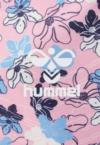 Logo blanc Hummel sur tissu rose avec un motif floral bleu, noir et blanc en arrière-plan.