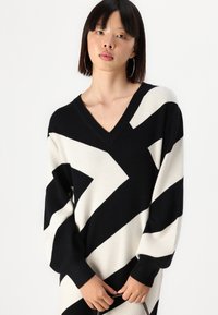 Pull en V avec de larges rayures en zigzag noir et crème, manches longues et poignets côtelés pour plus de texture. Tissu doux avec une coupe décontractée.