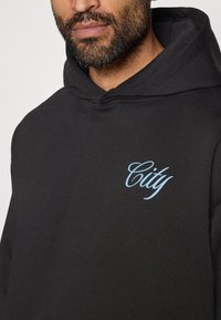 Mustmust suursuur musta tõmblīga, millel on sinine käekirjaline "Linn" embleem vasakul rinnakorvil. Materjal tundub pehme; disain hõlmab avarat kapuutsi.