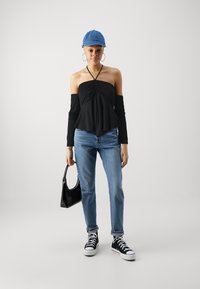 Zwart off-shoulder top met lange mouwen, gedrapeerd ontwerp. Lichtblauwe jeans, zwarte high-top sneakers en een blauwe denim pet. Zwart handtasaccessoire.