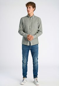 Camicia grigia chiara con bottoni, con tasca sul petto, indossata con jeans blu e sneakers bianche. Tessuto morbido, vestibilità su misura, stile casual.