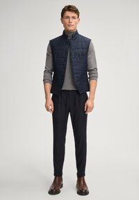 Granatowa pikowana kamizelka z wysokim kołnierzem, zapinana na guziki, zestawiona z szarym swetrem i ciemnymi spodniami. Brązowe skórzane buty dopełniają strój.