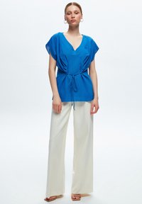 Blusa blu con scollo a V, maniche corte e un cordoncino in vita, abbinata a pantaloni larghi color crema e sandali beige.