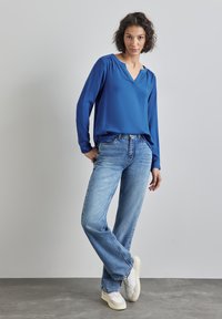 Blusa blu a maniche lunghe con scollo a V e spalle arricciate, abbinata a jeans larghi blu chiaro e sneakers bianche. Tessuto liscio in tutto.