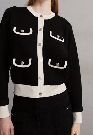 Frau trägt eine schwarze Strickjacke mit weißem Besatz, silbernen Knöpfen und vier dekorativen Vorderentaschen, kombiniert mit schwarzen, strukturierten Hosen.
