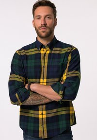 Langærmet ternet skjorte i navy, grøn og gul. Har krave med knapper, brystlomme og oprullede ærmer. Blødt stof med rutemønster.