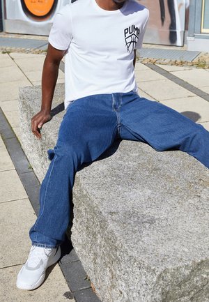 Homme portant un t-shirt blanc Puma, un jean bleu et des baskets blanches, assis sur un grand banc en pierre à l'extérieur à côté d'un mur de graffiti.