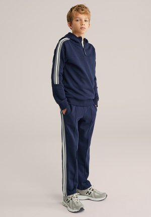 Tengerészkék melegítőfelső fél cipzárral, fehér csíkos díszítéssel, hozzáillő nadrággal; szürke sportcipővel párosítva. Sima anyagtextúra.