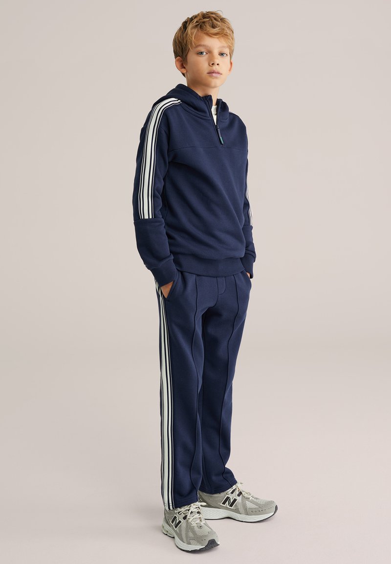 Tumši zils sporta tērps ar pus rāvējslēdzēja puloveri, baltām svītrām un atbilstošām biksēm; komplektā ar pelēkām sporta kurpēm. Gluda auduma tekstūra.