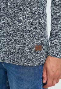 Pull en maille cable marine et blanc avec un motif texturé, des poignets côtelés et une petite étiquette en cuir brun au bas.