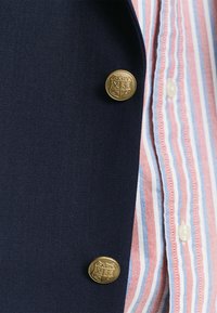 Blazer blu navy in tessuto testurizzato, con un bottone con emblema dorato. Sotto, una camicia a righe nei colori rosso, bianco e blu con un bottone bianco.
