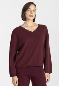 Bordowy sweter w serek z długimi rękawami, miękka faktura, luźny krój i ściągacz na dole, w połączeniu z dopasowanymi spodniami dla spójnego wyglądu.