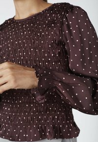 Nærbillede af en person iført en mørk lilla bluse med lyserøde prikker, som har smocket bærestykke og puffede ærmer med flæser.