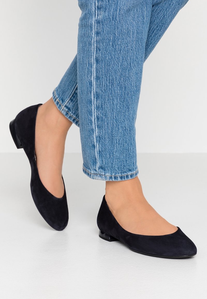 Femme portant un jean bleu et des ballerines noires en suède à talons bas et bouts pointus, debout sur un sol blanc.