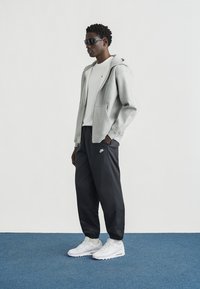 Grijze hoodie met rits, wit t-shirt, zwarte trainingsbroek met logo en witte sneakers. Model draagt een zonnebril en staat op een blauw tapijt.